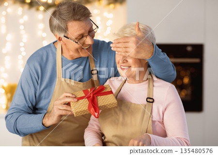 Happy Aged Man Suprising Wife With Christmas Gift Covering Her Eyes Standing In Modern Kitchen At Home. Winter Holidays Happiness And Celebration. New Year Present Concept 135480576