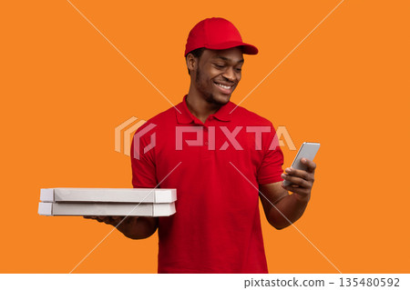 Fast Service And Best Food Delivery. Smiling black male courier wearing red uniform and cap holding pizza boxes from restaurant and using smartphone, isolated on orange studio background 135480592
