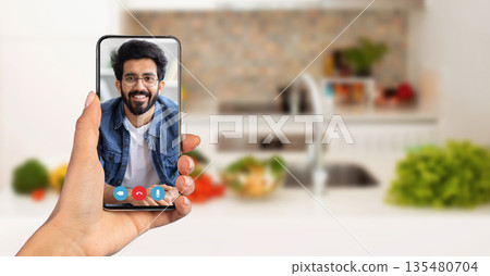 A person holds a smartphone displaying a smiling face during an online call. The background features a well-lit kitchen with fresh vegetables, capturing a moment of connection from home. A person holds a smartphone displaying a smiling face during an online call. The background features a well-lit kitchen with fresh vegetables, capturing a moment of connection from home. 135480704