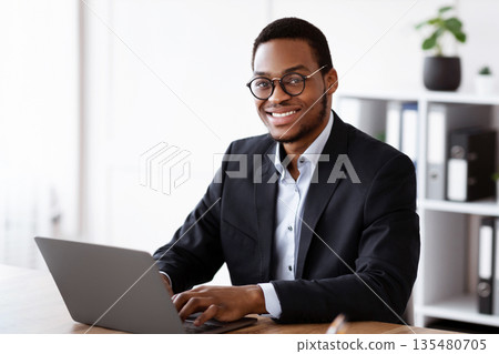 Cheerful african american businessman working with modern laptop in office, copy space. Happy young black manager in suit and glasses working online, typing emails or chatting with clients Cheerful african american businessman working with modern laptop in office, copy space. Happy young black manager in suit and glasses working online, typing emails or chatting with clients 135480705