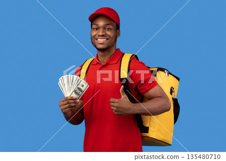 Payment For Delivery. Smiling african american male courier in red cap and uniform holding money, wearing yellow thermo backpack and showing thumbs up gesture, standing isolated over blue background Payment For Delivery. Smiling african american male courier in red cap and uniform holding money, wearing yellow thermo backpack and showing thumbs up gesture, standing isolated over blue background 135480710