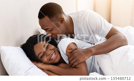 Happy loving african-american couple relaxing in bed at home, enjoying togetherness and tenderness in love, copy space. Cheerful black family man and woman in love having fun in bedroom 135480712