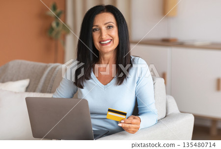 Easy Payment And Online Transaction. Smiling mature woman ordering on internet, using laptop, showing plastic credit card for shopping in virtual store. Lady sitting on sofa at home, looking at camera 135480714