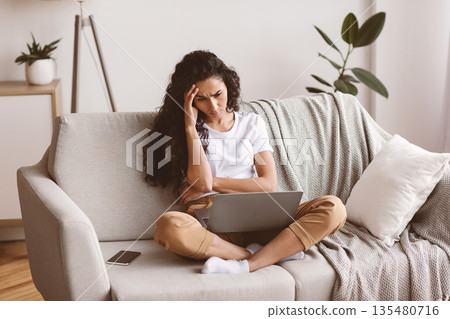 Upset young jobless woman sitting on couch with laptop on her lap, touching her head and looking at screen, home interior. Thoughtful curly dark-haired lady having financial difficulties, copy space Upset young jobless woman sitting on couch with laptop on her lap, touching her head and looking at screen, home interior. Thoughtful curly dark-haired lady having financial difficulties, copy space 135480716