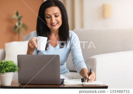 Home Office. Portrait of smiling middle-aged woman writing in her notebook, working on laptop, taking notes, holding cup and drinking coffee, sitting on the couch at table with personal computer Home Office. Portrait of smiling middle-aged woman writing in her notebook, working on laptop, taking notes, holding cup and drinking coffee, sitting on the couch at table with personal computer 135480731