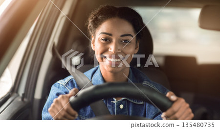 Happy woman driving car and smiling going to work or on road trip. Cute young positive happy african american female with safety belt holds steering wheel looking through the windshield, empty space Happy woman driving car and smiling going to work or on road trip. Cute young positive happy african american female with safety belt holds steering wheel looking through the windshield, empty space 135480735