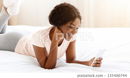 Relaxed african american young lady laying on bed with smartphone, chatting with friends via social media or reading blog, using nice applications, resting on weekend at home, copy space Relaxed african american young lady laying on bed with smartphone, chatting with friends via social media or reading blog, using nice applications, resting on weekend at home, copy space 135480744