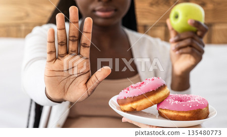 Healthy Eating During Pregnancy. Black Pregnant Lady Refusing Unhealthy Donuts And Choosing Apple For Snack, Showing Stop Gesture With Palm, Sitting On Bed At Home, Closeup Shot With Selective Focus Healthy Eating During Pregnancy. Black Pregnant Lady Refusing Unhealthy Donuts And Choosing Apple For Snack, Showing Stop Gesture With Palm, Sitting On Bed At Home, Closeup Shot With Selective Focus 135480773