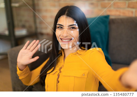 A woman smiles and waves while using facial recognition technology for identification. This scene captures the integration of advanced tech in everyday activities, reflecting the new normal. A woman smiles and waves while using facial recognition technology for identification. This scene captures the integration of advanced tech in everyday activities, reflecting the new normal. 135480777