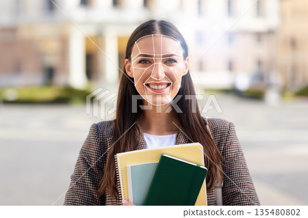 A young woman stands in a city plaza holding notebooks and smiles as facial recognition technology analyzes her features. The scene reflects the integration of modern tech in daily life. A young woman stands in a city plaza holding notebooks and smiles as facial recognition technology analyzes her features. The scene reflects the integration of modern tech in daily life. 135480802