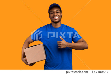 Best Delivery Service Concept. Smiling black male courier wearing blue cap and uniform holding cardboard box and showing thumbs up gesture standing isolated over yellow orange studio background Best Delivery Service Concept. Smiling black male courier wearing blue cap and uniform holding cardboard box and showing thumbs up gesture standing isolated over yellow orange studio background 135480867