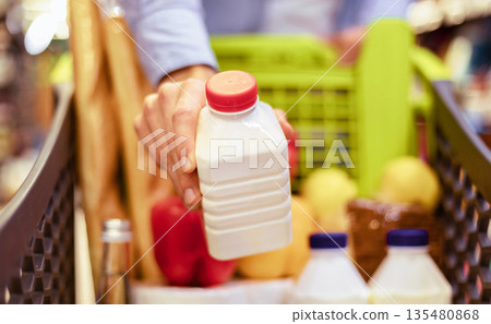 Grocery Shopping. Closeup of unrecognizable male customer holding bottle of milk or yoghurt and putting it in trolley cart full of food. Young man buying healthy dairy products in the mall Grocery Shopping. Closeup of unrecognizable male customer holding bottle of milk or yoghurt and putting it in trolley cart full of food. Young man buying healthy dairy products in the mall 135480868