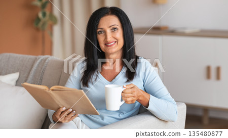 Spending Time At Home. Portrait of mature lady reading book and holding cup of coffee, sitting on the couch in living room in her apartment, enjoying weekend. Woman looking at camera Spending Time At Home. Portrait of mature lady reading book and holding cup of coffee, sitting on the couch in living room in her apartment, enjoying weekend. Woman looking at camera 135480877