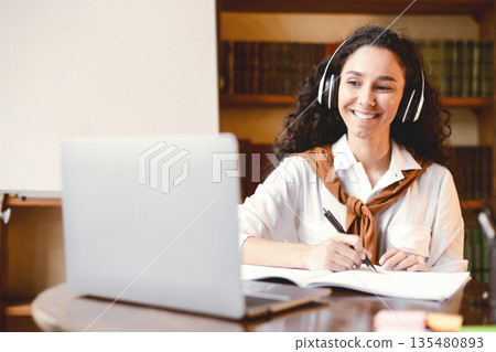 Distance Education. Smiling woman in wireless headset sitting at desk, using laptop and writing in notebook. Lady watching tutorial, lecture or webinar, studying online at home. E-learning Distance Education. Smiling woman in wireless headset sitting at desk, using laptop and writing in notebook. Lady watching tutorial, lecture or webinar, studying online at home. E-learning 135480893