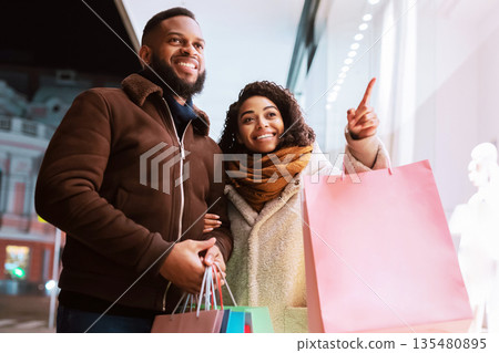Sales And Discount. Cheerful curly black woman pointing finger at mall window, indicating showcase, walking with her man in the evening near shopping center, holding colorful bags with purchases Sales And Discount. Cheerful curly black woman pointing finger at mall window, indicating showcase, walking with her man in the evening near shopping center, holding colorful bags with purchases 135480895