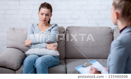 Upset young lady suffering from PTSD, sitting on couch at counselor's office and hugging pillow. Depressed female patient consulting with her psychologist, receiving treatment of mood disorder 135480896