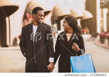 Man and woman with shopping bags talking and smiling, walking down the street after Black Friday and season sale. Millenial african american family holds bags with purchases and look at each other Man and woman with shopping bags talking and smiling, walking down the street after Black Friday and season sale. Millenial african american family holds bags with purchases and look at each other 135480901