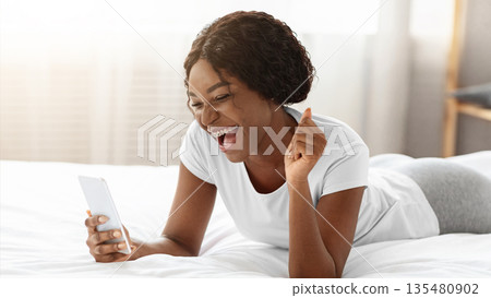 Emotional african american woman looking at smartphone screen, having good news, laying on bed at home, free space. Happy black lady with mobile phone clenching fist, playing mobile games Emotional african american woman looking at smartphone screen, having good news, laying on bed at home, free space. Happy black lady with mobile phone clenching fist, playing mobile games 135480902