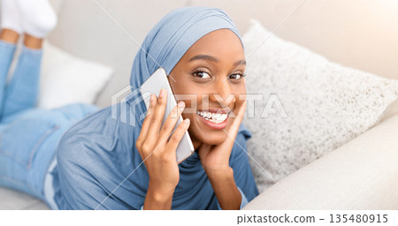 Attractive young Muslim woman in hijab chatting on cellphone, having pleasant talk with friend while lying on sofa at home. Modern technologies and communication concept 135480915