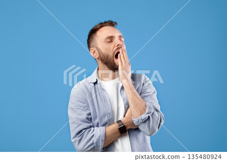 Bored young guy yawning, covering open mouth with hand over blue studio background. Tired millennial man feeling sleepy, suffering from insomnia, not interested in something Bored young guy yawning, covering open mouth with hand over blue studio background. Tired millennial man feeling sleepy, suffering from insomnia, not interested in something 135480924