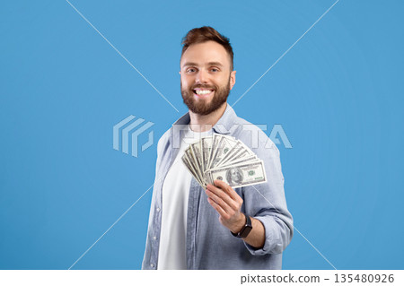 Happy young man posing with lots of money, holding dollar fan over blue studio background. Lucky millennial guy showing casino or lottery win, sharing financial success, offering profit Happy young man posing with lots of money, holding dollar fan over blue studio background. Lucky millennial guy showing casino or lottery win, sharing financial success, offering profit 135480926