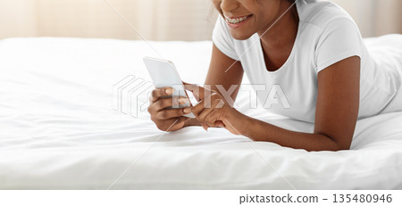 Happy black lady laying on bed at home, using mobile phone and smiling, copy space. African american woman spending weekend at home, chatting with friends or checking social media on smartphone Happy black lady laying on bed at home, using mobile phone and smiling, copy space. African american woman spending weekend at home, chatting with friends or checking social media on smartphone 135480946