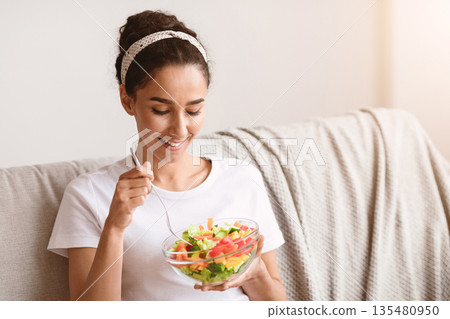 Healthy lifestyle, diet concept. Smiling millennial woman eating fresh salad vegetables, sitting on sofa in living room, copy space. Closeup of happy young lady enjoying healthy food Healthy lifestyle, diet concept. Smiling millennial woman eating fresh salad vegetables, sitting on sofa in living room, copy space. Closeup of happy young lady enjoying healthy food 135480950
