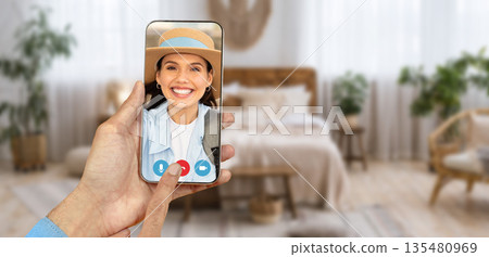 A person is using a smartphone to participate in a video call from their cozy living room. The screen displays a smiling face, highlighting the connectivity of online communication from home. 135480969
