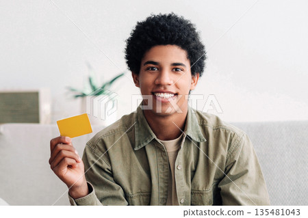 Internet shopping concept. Positive black teenager showing credit card in front of laptop, ordering stuff online from home. Funky African American teen guy purchasing on web indoors, panorama 135481043