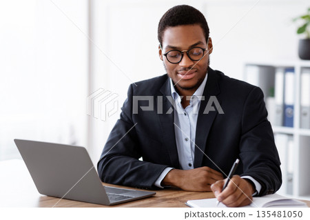 Enthusiastic black businessman planning his day in office, sitting in front of modern laptop and writing in notebook, copy space. Young african american manager having online training, taking notes Enthusiastic black businessman planning his day in office, sitting in front of modern laptop and writing in notebook, copy space. Young african american manager having online training, taking notes 135481050
