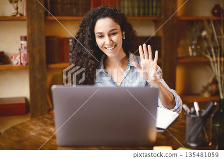 Teleconference And Telecommuting. Portrait of smiling woman sitting at desk, having video call on laptop, waving to webcam. Distance Education. Cheerful lady wearing headphones at virtual meeting Teleconference And Telecommuting. Portrait of smiling woman sitting at desk, having video call on laptop, waving to webcam. Distance Education. Cheerful lady wearing headphones at virtual meeting 135481059