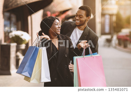 Fun and shopping together on black friday and final sale. Happy african american young guy and lady in coats enjoying walk, look at each other and hold many colorful bags with purchase, copy space 135481071