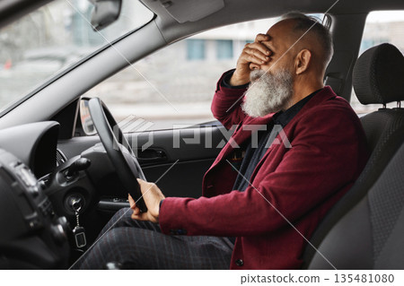 Exhausetd aged grey-haired man having headache while driving car, touching his forehead, side view, shot inside auto, copy space. Elderly businessman having financial crisis or got stuck in traffic 135481080