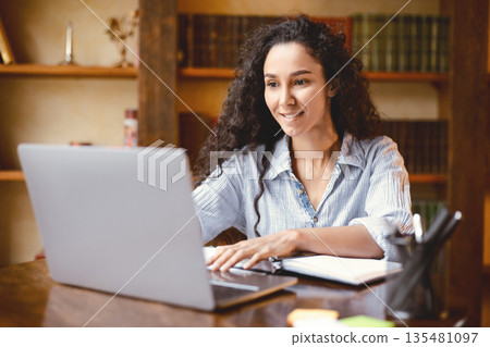 Doing Project. Portrait of curly young woman using laptop computer and typing on keyboard, writer sitting at table. Female manager working remotely or studying online at home, telecommuting Doing Project. Portrait of curly young woman using laptop computer and typing on keyboard, writer sitting at table. Female manager working remotely or studying online at home, telecommuting 135481097