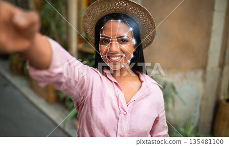 A person is engaging with innovative facial recognition technology outdoors. The individual is smiling while showcasing a digital overlay highlighting facial features in a vibrant urban setting. 135481100