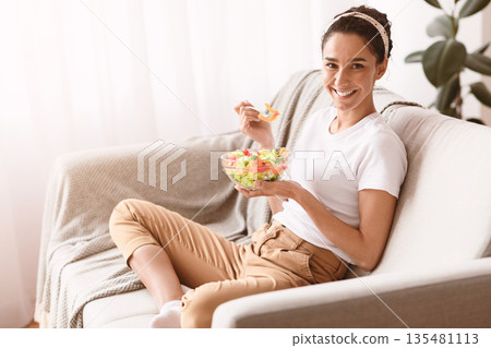 Diet, nutrition, healthy lifestyle concept. Cheerful young lady holding bowl with veggies salad, sitting on couch in living room, enjoying her healthy snack and smiling at camera, copy space Diet, nutrition, healthy lifestyle concept. Cheerful young lady holding bowl with veggies salad, sitting on couch in living room, enjoying her healthy snack and smiling at camera, copy space 135481113