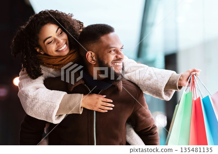 Healthy Relationship. Smiling african american man giving piggyback ride to his happy woman who holding shopping bags, couple looking at shop window, walking in the evening near center 135481135