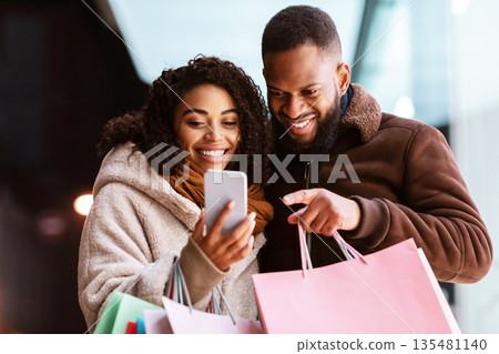 Sales, Offers And Discount. Smiling african american family of two using mobile phone, guy pointing at gadget, couple standing outdoors in the evening and holding colorful shopping bags 135481140