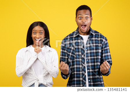 Wow, it is win. Excited millennial african american couple in love with wide open eyes and mouths, expresses emotions of victory and look at camera, isolated on yellow background, studio shot 135481141