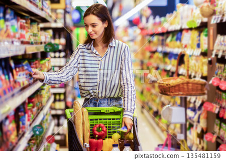 At The Supermarket. Portrait Of Young Woman Standing With Trolley Cart Between Aisles In Grocery Store. Cheerful Consumer Buying Essentials In Local Shop, Taking Products From Shelf 135481159