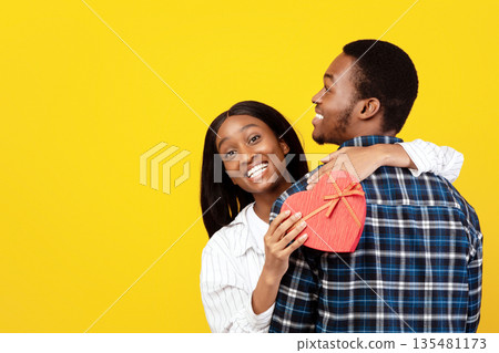 Happiness together, anniversary, Valentine's day and love. Smiling young african american wife hugging husband and holding box in heart shaped, isolated on yellow background, studio shot, free space Happiness together, anniversary, Valentine's day and love. Smiling young african american wife hugging husband and holding box in heart shaped, isolated on yellow background, studio shot, free space 135481173