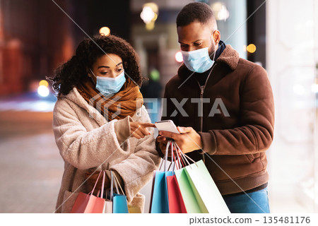 Retail, Technology, Protection And Pandemic Concept. Portrait of black couple wearing medical masks, using mobile phone, lady pointing at screen, holding shopper bags, walking in the evening Retail, Technology, Protection And Pandemic Concept. Portrait of black couple wearing medical masks, using mobile phone, lady pointing at screen, holding shopper bags, walking in the evening 135481176