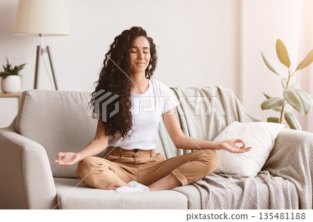 Relaxation, stress relief concept. Peaceful brunette lady with long curly hair sitting in lotus position on couch in living room, meditating, copy space. Positive young woman enjoying weekend at home Relaxation, stress relief concept. Peaceful brunette lady with long curly hair sitting in lotus position on couch in living room, meditating, copy space. Positive young woman enjoying weekend at home 135481188