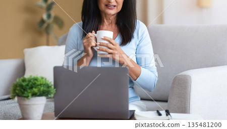 Technology And People. Attractive Mature Lady With Long Dark Hair Working Remotely On Laptop At Home Office, Enjoying Morning Coffee, Woman Sitting On The Couch With Computer On Desk 135481200