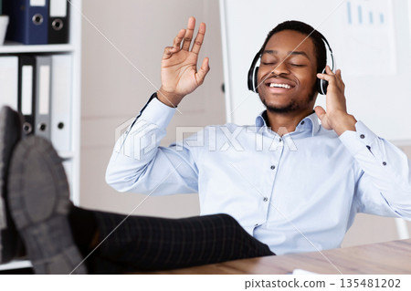 Joyful black manager leaning back with legs on workdesk and listening to music with closed eyes in his office, singing songs, using wireless headset, copy space. Stress relief at job concept Joyful black manager leaning back with legs on workdesk and listening to music with closed eyes in his office, singing songs, using wireless headset, copy space. Stress relief at job concept 135481202