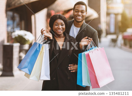 Season sale, consumerism, Black friday and family walk. Smiling young african american couple hugs, lady holds a lot of colored bags with gifts for holidays near shopping mall, outdoor, free space 135481211