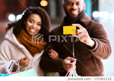 Consumerism And Relationship. Portrait of smiling black man showing debit credit card to camera, woman embracing him and holding shopping bags, family near mall in the evening, blurred background Consumerism And Relationship. Portrait of smiling black man showing debit credit card to camera, woman embracing him and holding shopping bags, family near mall in the evening, blurred background 135481218