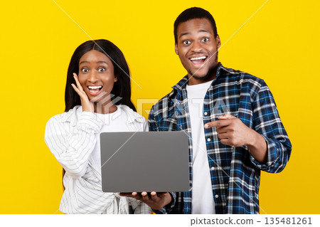 Amazing offer, good site and celebrating victory. Surprised cheerful millennial african american husband and wife hold laptop, man points finger at gadget, isolated on yellow background, studio shot Amazing offer, good site and celebrating victory. Surprised cheerful millennial african american husband and wife hold laptop, man points finger at gadget, isolated on yellow background, studio shot 135481261