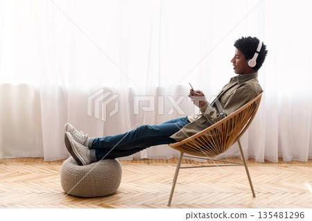 Black teenager relaxing in modern armchair, enjoying music on smartphone at home, banner design with copy space. African American youth in headphones listening to his favorite song Black teenager relaxing in modern armchair, enjoying music on smartphone at home, banner design with copy space. African American youth in headphones listening to his favorite song 135481296
