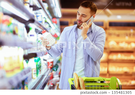Grocery Shopping. Portrait of male customer holding bottle of milk or yoghurt, talking on smartphone, asking wife about list of purchases, standing near fridge with dairy products Grocery Shopping. Portrait of male customer holding bottle of milk or yoghurt, talking on smartphone, asking wife about list of purchases, standing near fridge with dairy products 135481305